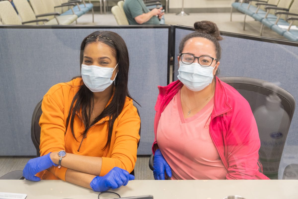Two women of colour smiling with masks on behind a check in desk.