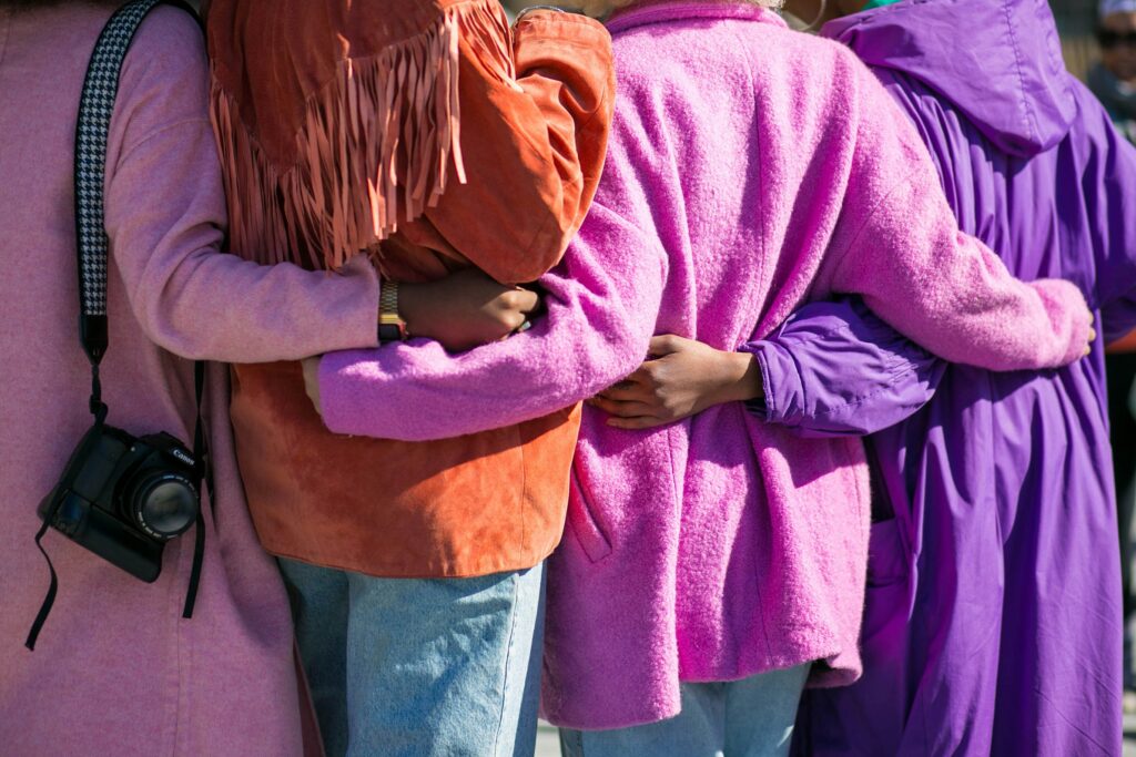 Photographed from behind, four women dressed in pink, purple and orange coats hold each other.